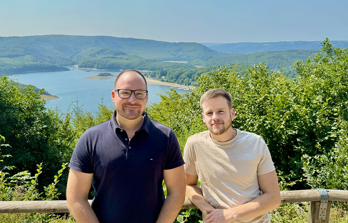 Zwei Personen an einem Aussichtspunkt mit See und bewaldeten Hügeln im Hintergrund